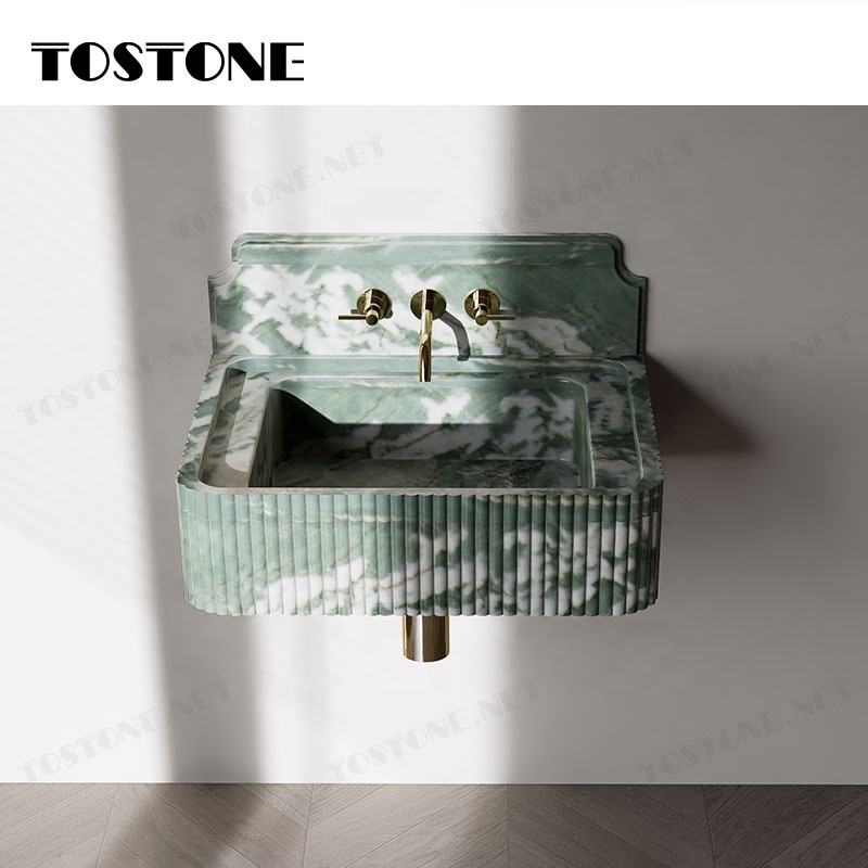 Bồn rửa mặt đá cẩm thạch xanh, vòi nước vàng, tường trắng, ánh sáng chiếu từ bên phải.