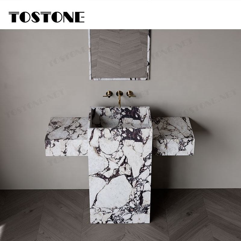 Bồn rửa mặt đá hoa cương trắng với vân đen, vòi nước vàng trên nền tường xám.