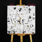Tấm gạch terrazzo vuông 600x600mm, nền trắng, chấm màu đen và nâu, trên giá vẽ gỗ.