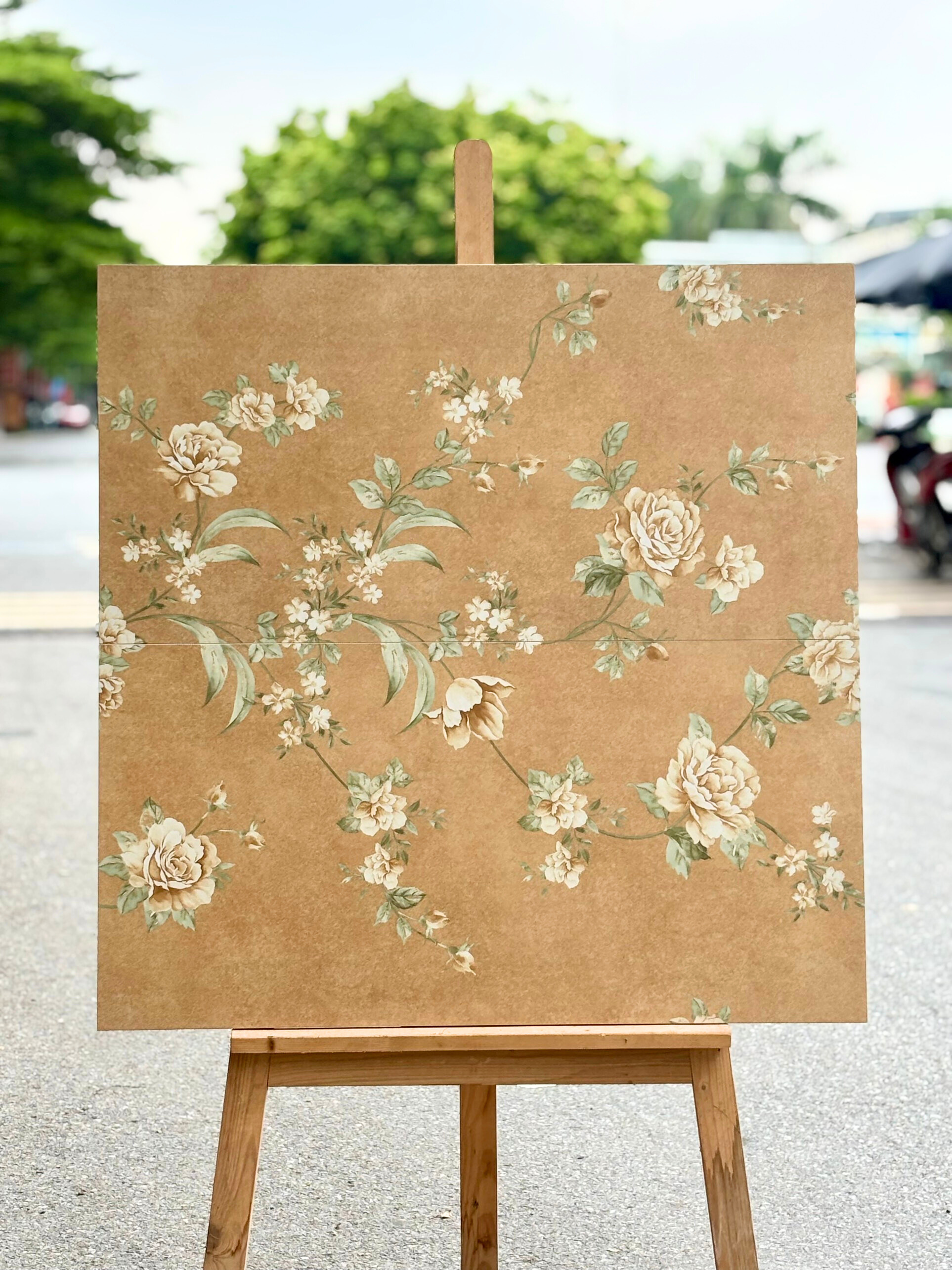 Tranh nền màu nâu nhạt với hoa trắng và lá xanh, đặt trên giá gỗ ngoài trời.