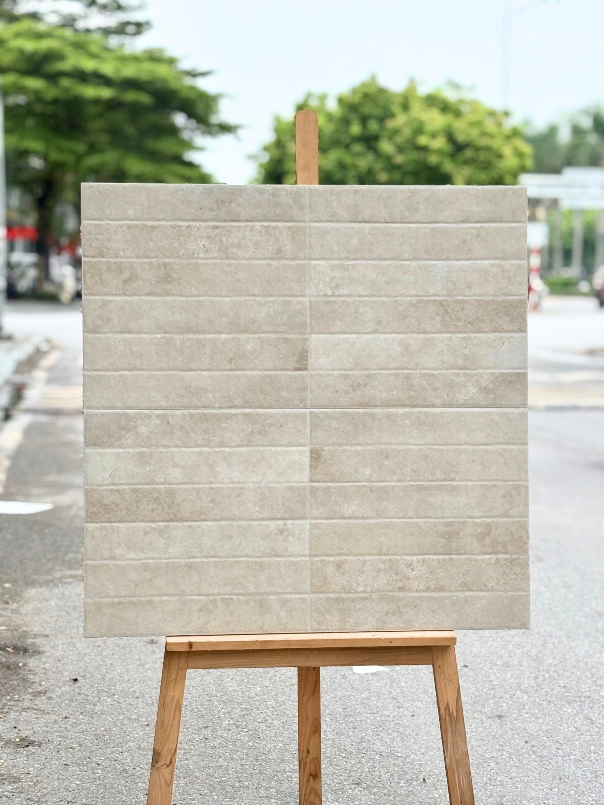Tấm gạch màu be vân đá trên giá gỗ giữa đường phố, nền là cây xanh nhạt nhòa.