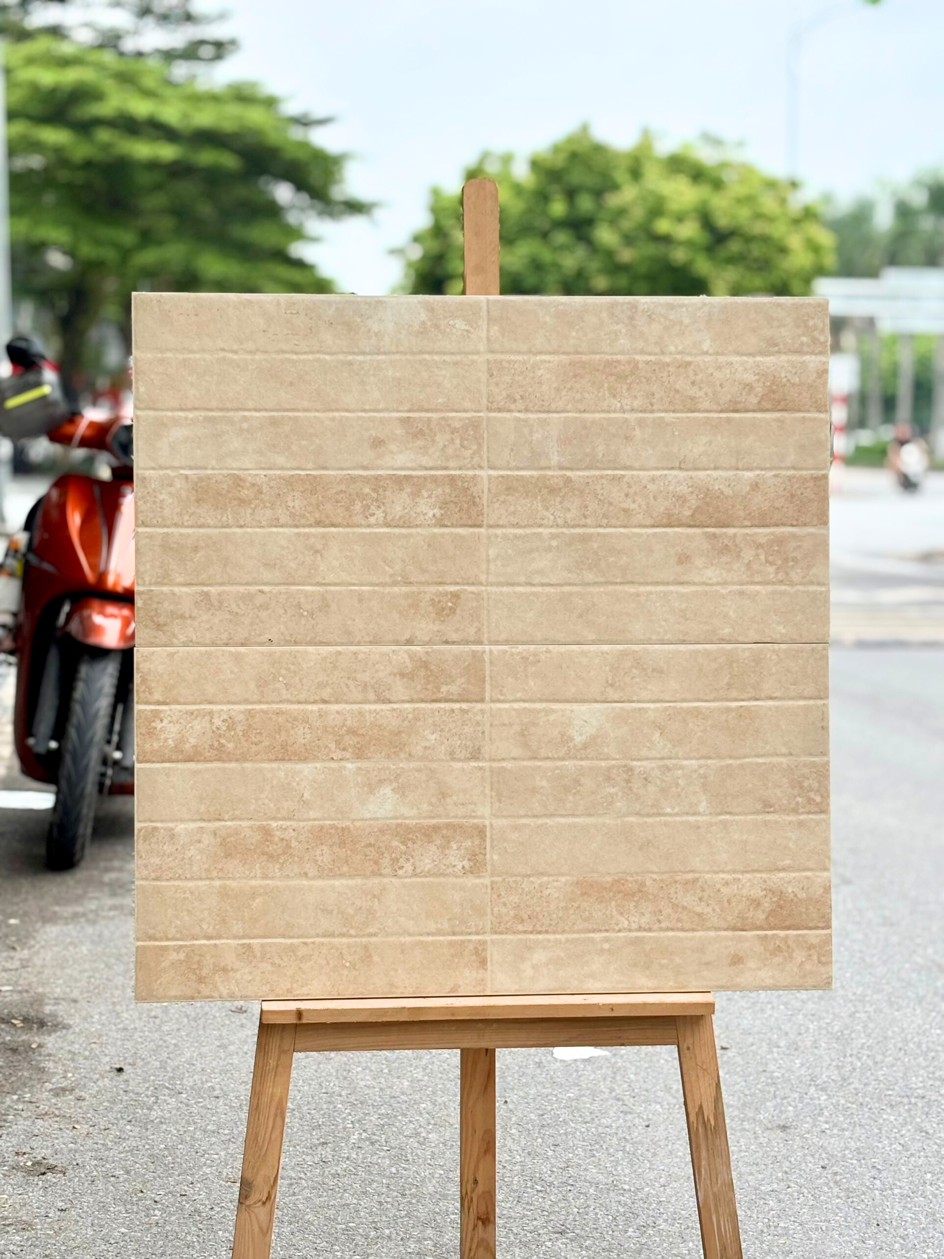 Tấm gạch màu be xếp trên giá gỗ ngoài trời, nền đường và cây xanh phía sau.