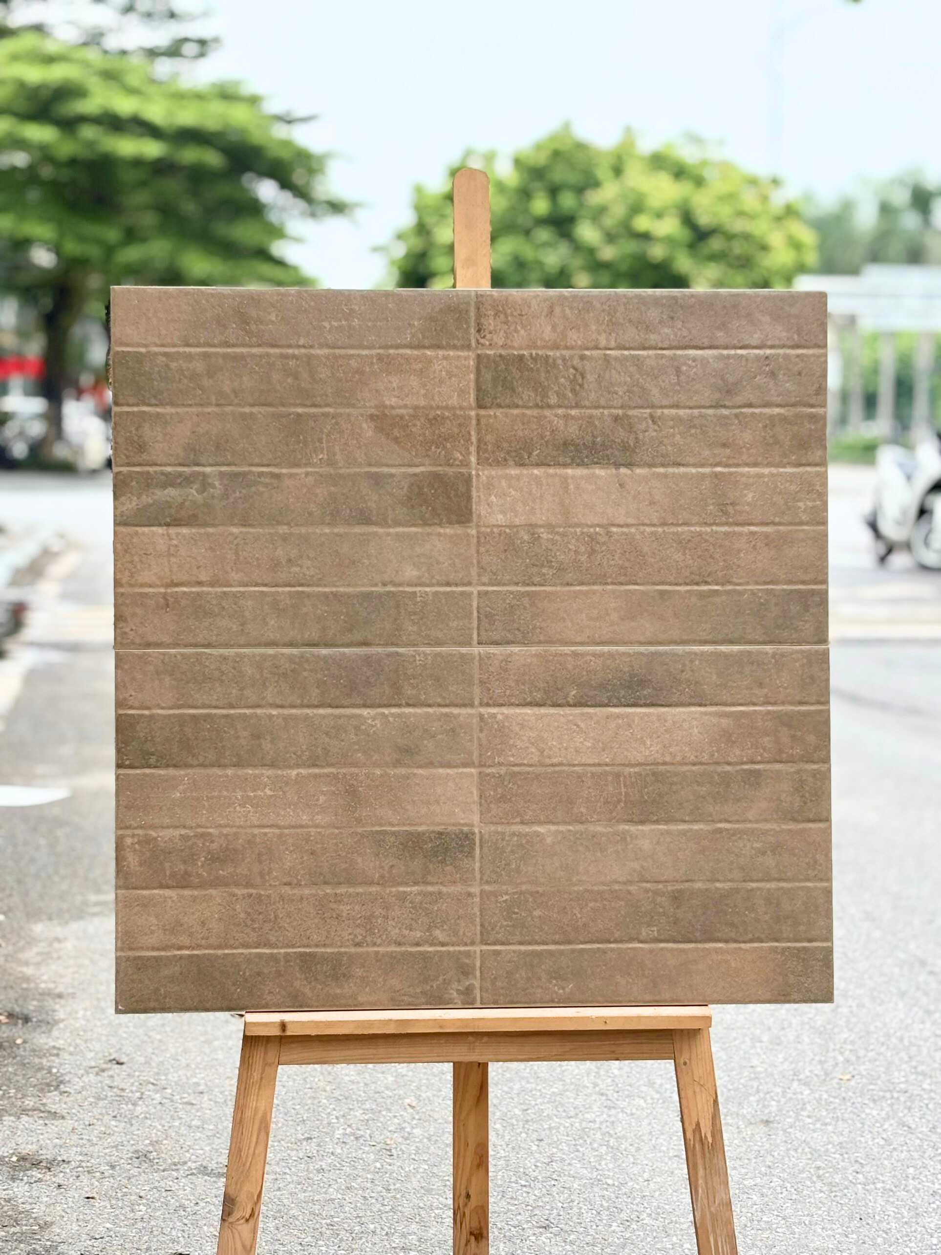Tấm bảng gạch nâu dựng trên giá gỗ, nền đường phố và cây xanh phía xa.