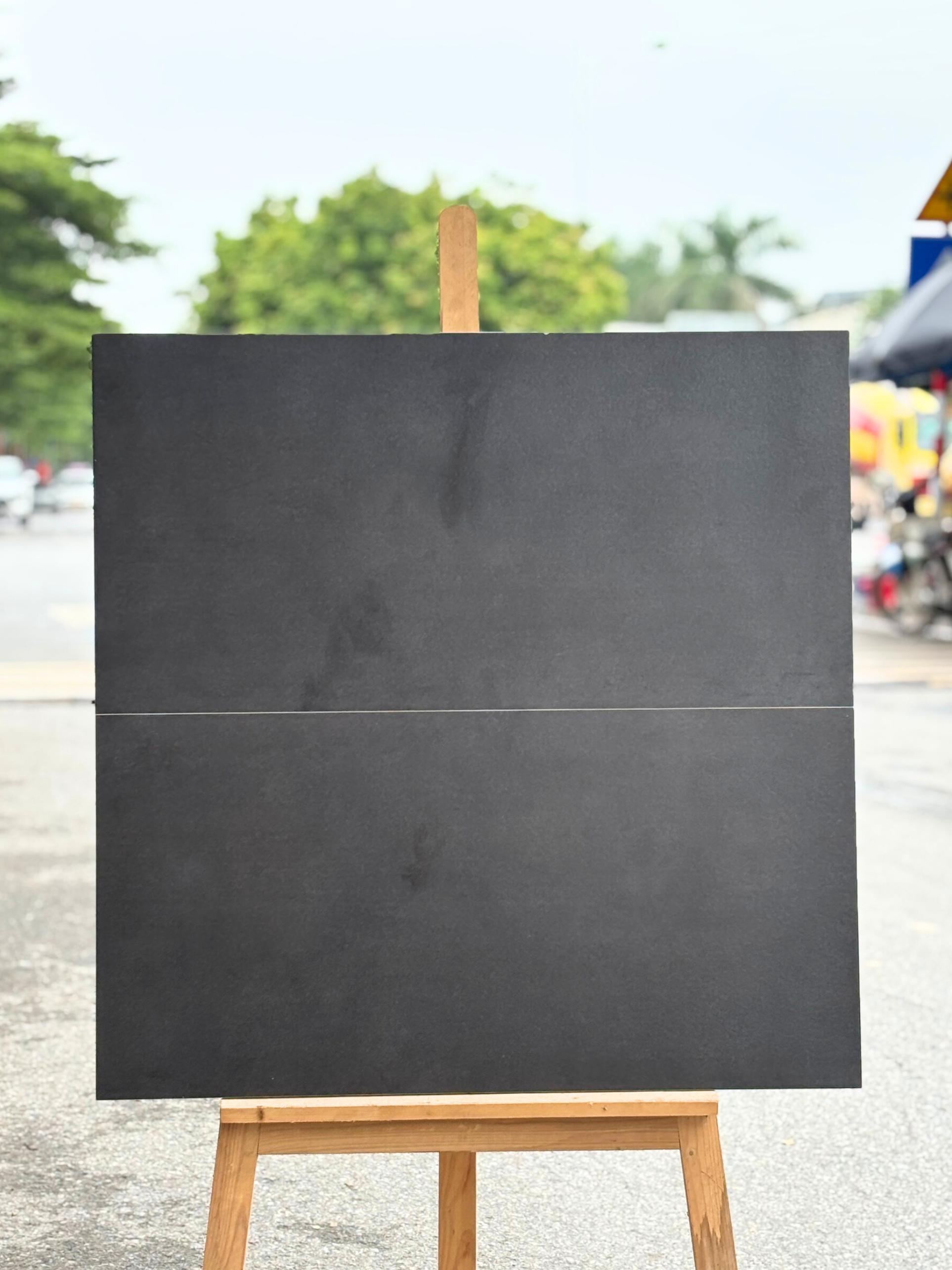 Tấm bảng đen trên giá vẽ gỗ, nền ngoài trời có cây xanh và lều chợ màu sắc.