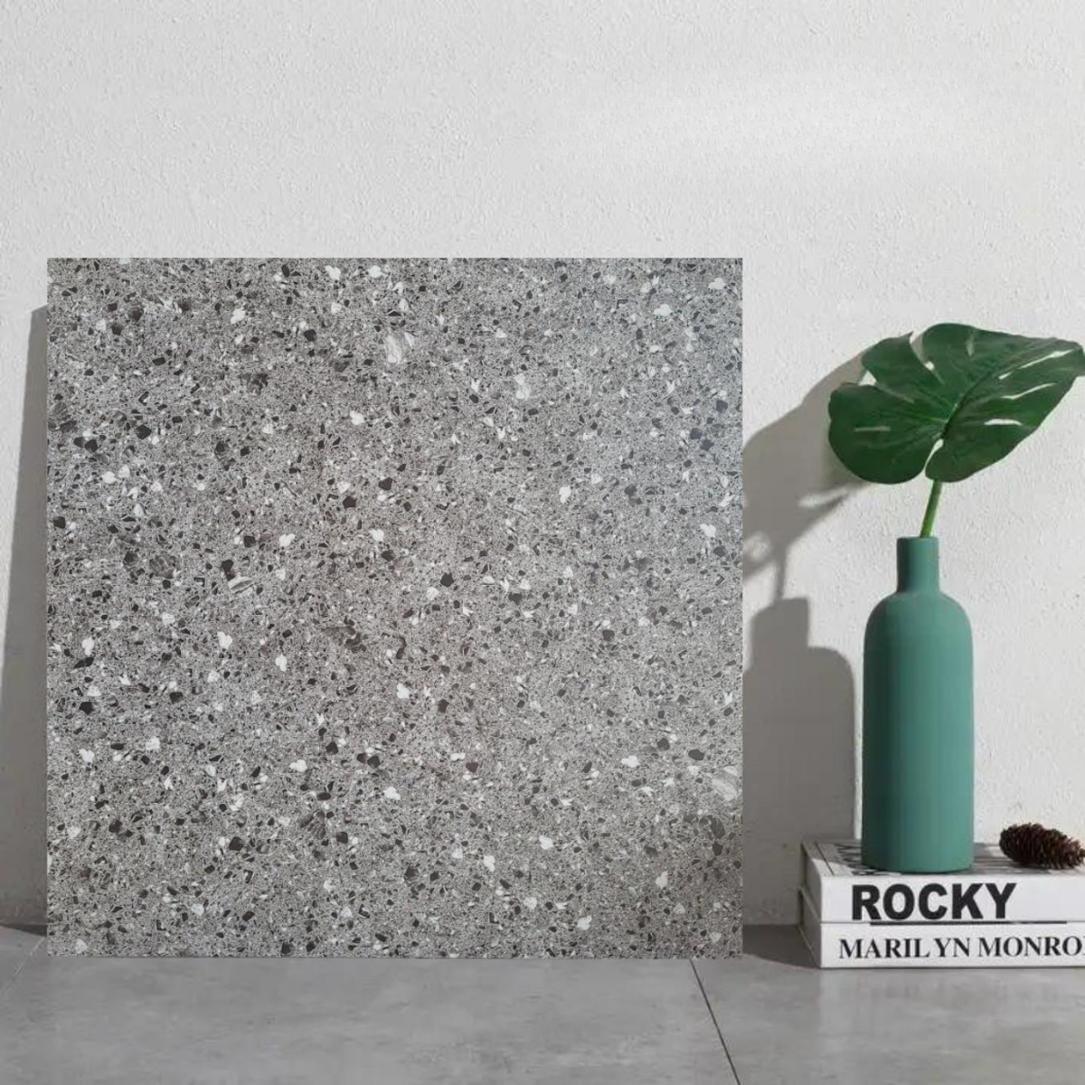 Tấm đá terrazzo xám, cạnh bình xanh với lá lớn, đặt trên sách trắng nền tường trắng.