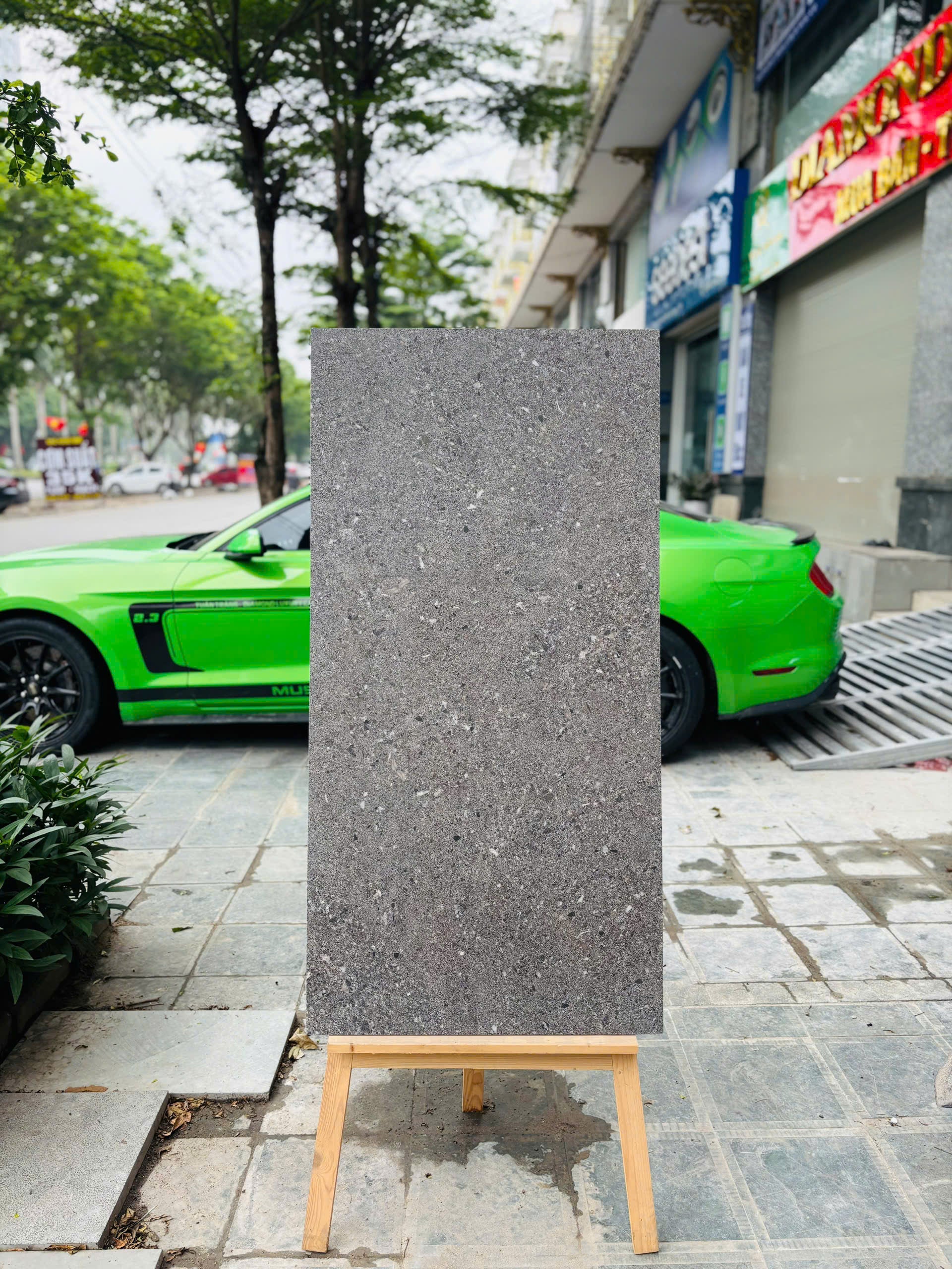 Tấm bảng đá xám đứng trên giá gỗ, nền là xe hơi màu xanh lá và hàng cây ven đường.